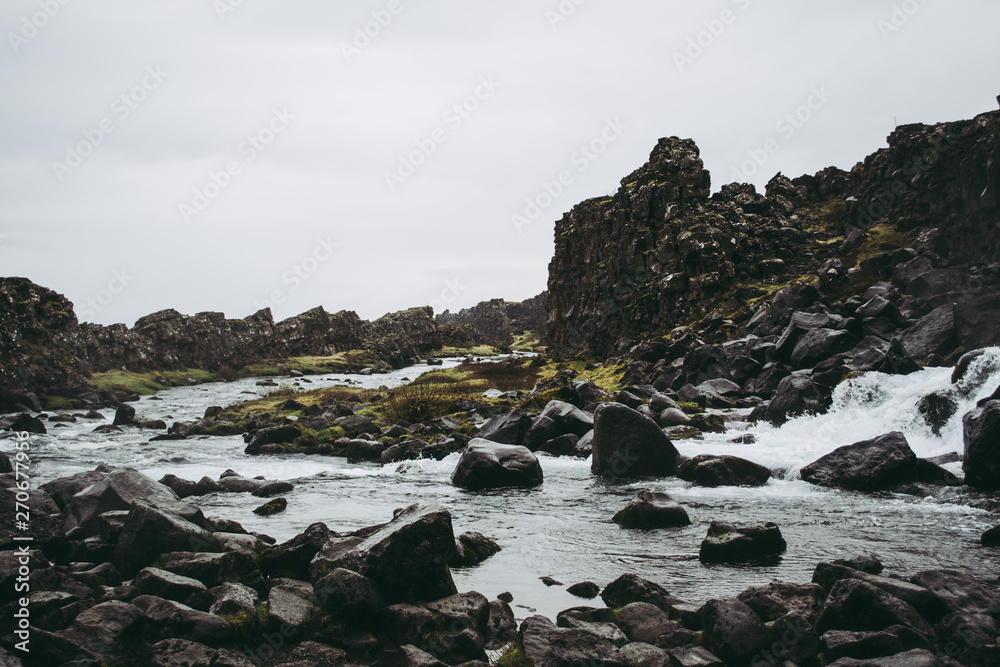 A mountain stream of dark cold water near the waterfall of Oxararfoss in Thingvellir National Park, Iceland. Green grass amidst the rocks beneath heavy gray sky