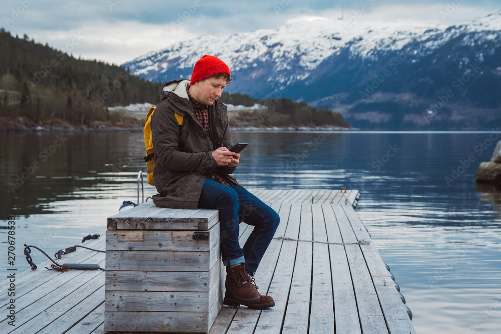 Traveler man looking into the phone. Tourist in yellow backpack sitting on a background of a mountain and a lake a wooden pier. Traveler walks, takes photo in mountains. Traveler man looking into the phone. Tourist in yellow backpack sitting on a background of a mountain and a lake a wooden pier. Traveler walks, takes photo in mountains.