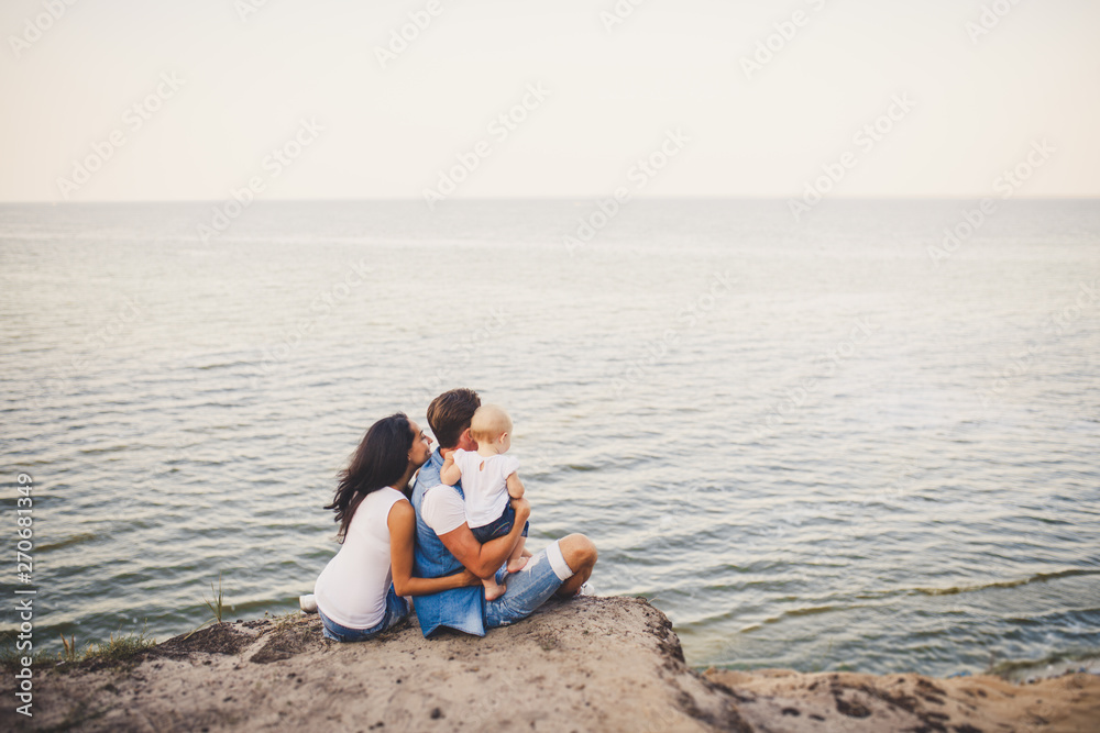 Theme family vacation with small child on the nature and sea. Mom, Dad and daughter of one year are sitting in embrace, girls in the hands of man, with his back at height of cliff overlooking the sea