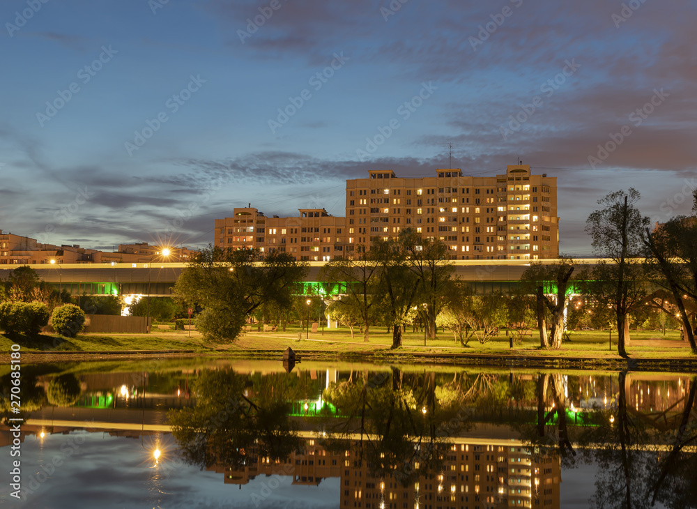 Evening cityscape. Lighted houses, paths in the city park, blurred track of the train reflected in the city pond against the bright evening sky