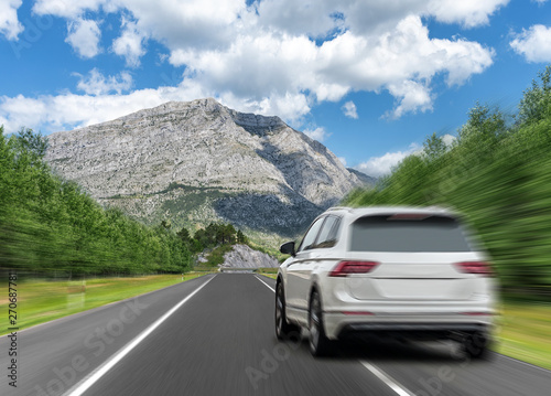 White car drives fast on the highway against the backdrop of a mountain range.