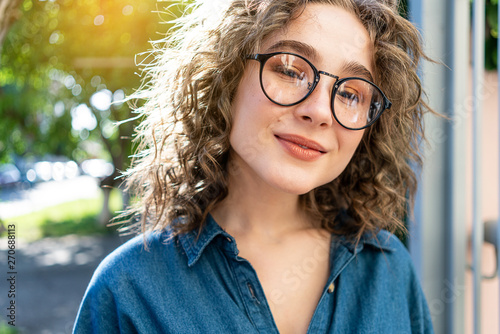 Outdoor close up portrait of young beautiful stylish happy smiling curly girl wearing sunglasses, posing in street. Sunny day light. Summer fashion concept. Copy, empty space for text