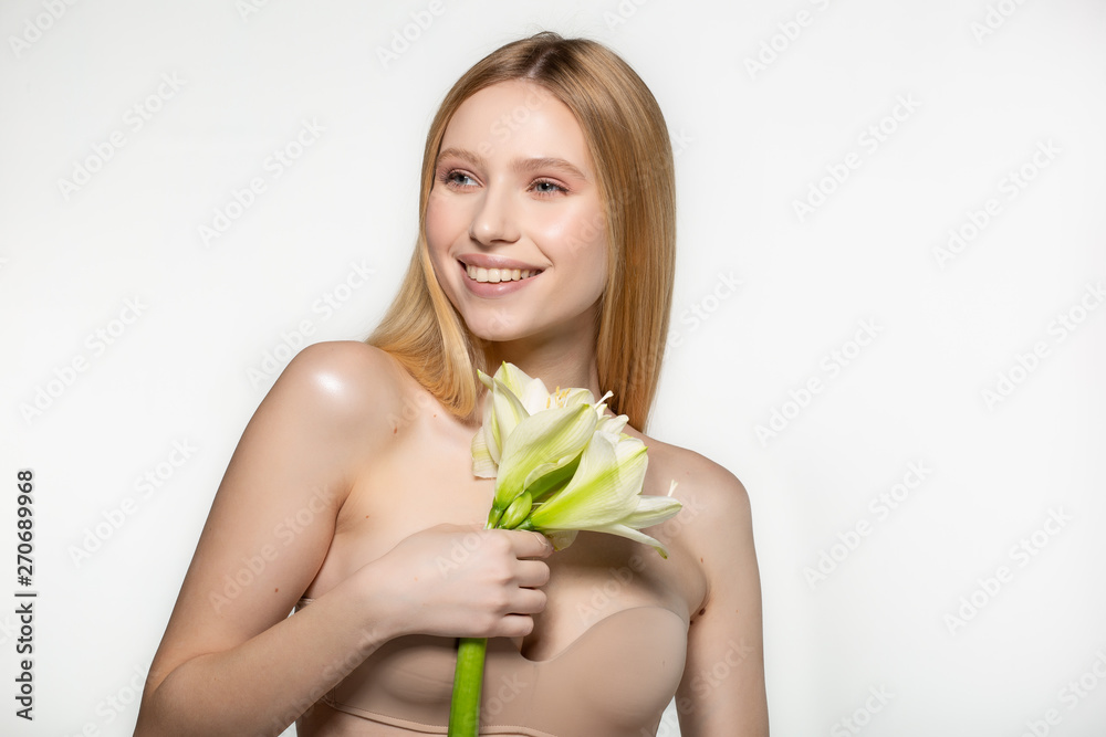 Laughing beautiful blonde girl posing with big white flower, looking away, half length portrait in the studio over grey background Laughing beautiful blonde girl posing with big white flower, looking away, half length portrait in the studio over grey background