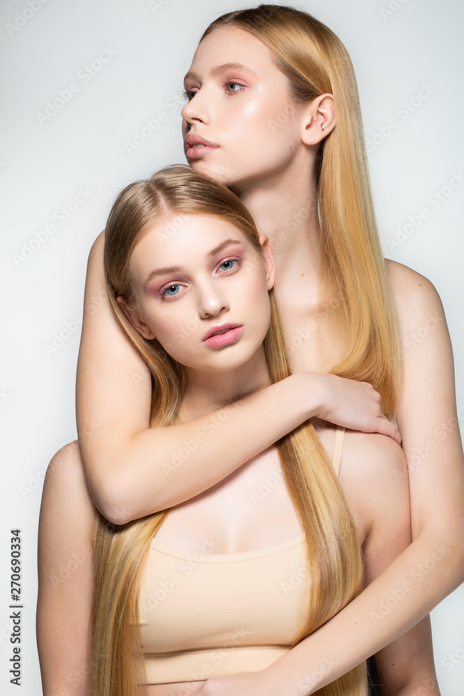 Two sexy attractive twins woman with blonde long hair posing in glamour makeup, standin behind each other, posing in the studio over grey background. Lesbian family couple Two sexy attractive twins woman with blonde long hair posing in glamour makeup, standin behind each other, posing in the studio over grey background. Lesbian family couple