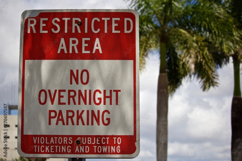 Restricted Area No overnight Parking Sign Red and white warning sign with palm trees and partly cloudy sky in background Restricted Area No overnight Parking Sign Red and white warning sign with palm trees and partly cloudy sky in background