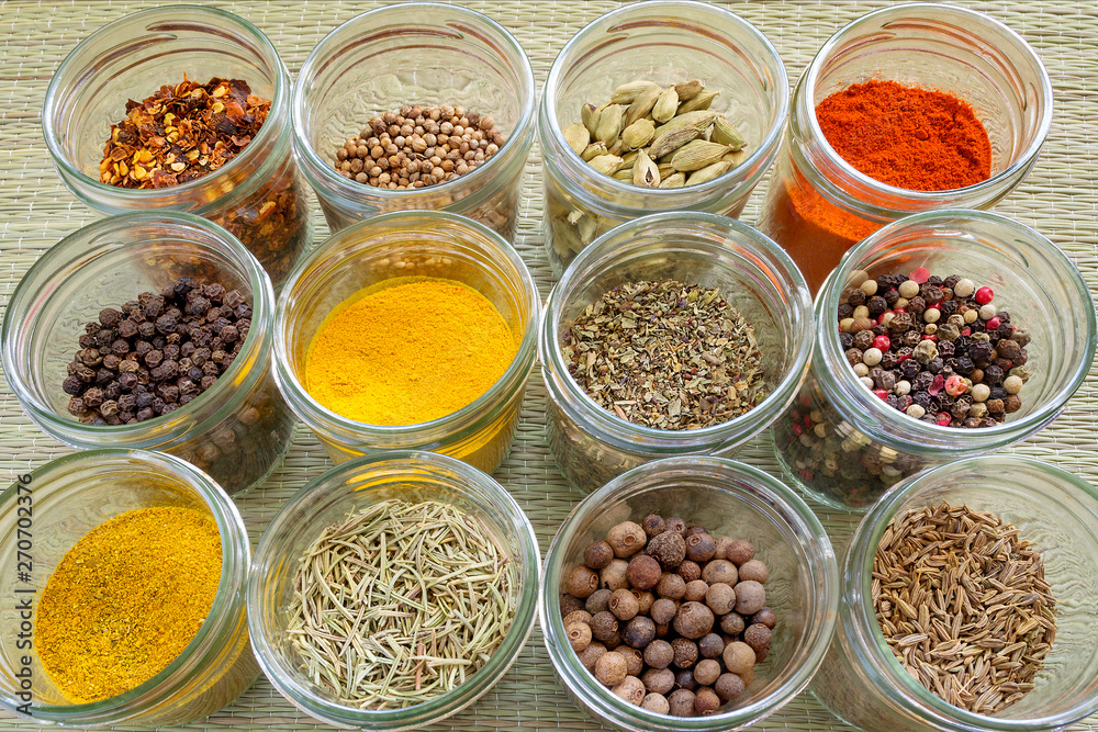 Twelve different spices and herb seasonings in a glass jars on a rough green mat made of natural plant fibers. Natural food spices and seasonings. Tasty eating. Twelve different spices and herb seasonings in a glass jars on a rough green mat made of natural plant fibers. Natural food spices and seasonings. Tasty eating.