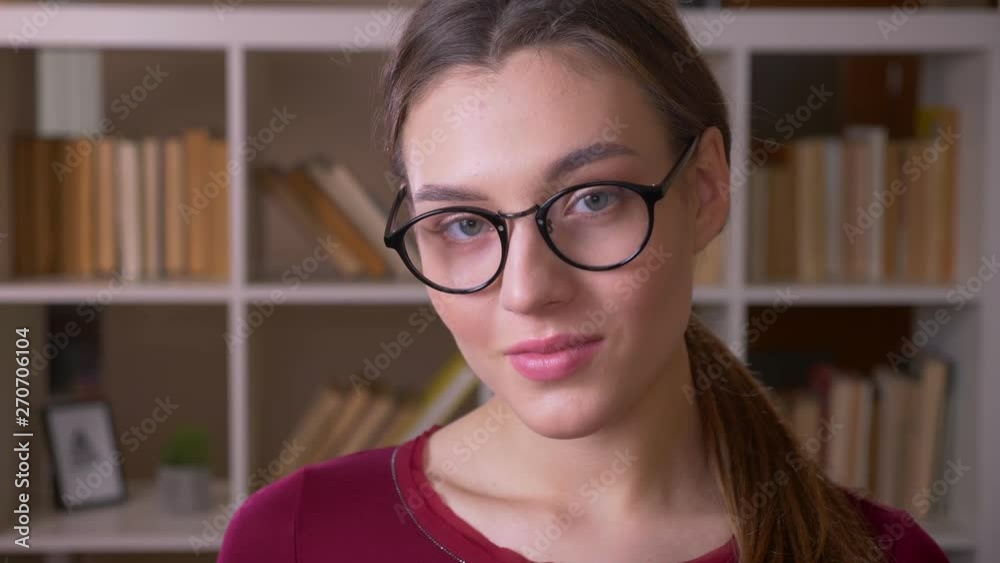 Portrait of young brunette female student with ponytail in eye glasses smiles with flirtation into camera in library. Portrait of young brunette female student with ponytail in eye glasses smiles with flirtation into camera in library.