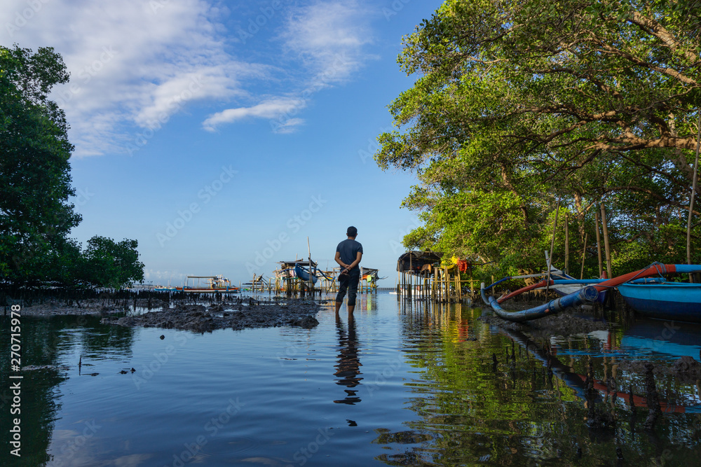 A man was standing in the middle of a mangrove forest whose sea water was receding. In front of him is a Balinese traditional boat view under the blue sky