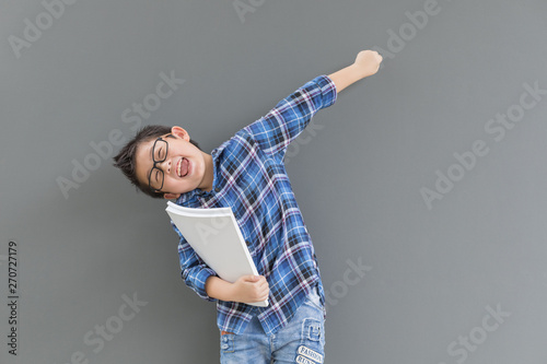 Asian boy raise hand up feels happiness when time to school and his hand hold the book against the gray wall