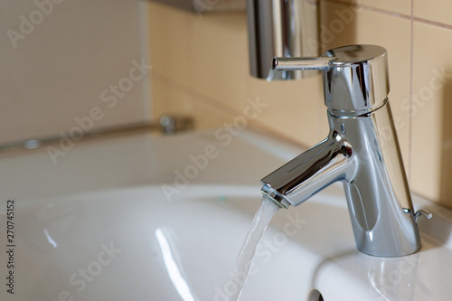 Water from the tap. A stream of clean water flows into the sink. Open chrome faucet washbasin. Warm tones.