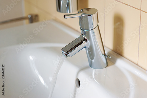 Water from the tap. A stream of clean water flows into the sink. Open chrome faucet washbasin. Warm tones.