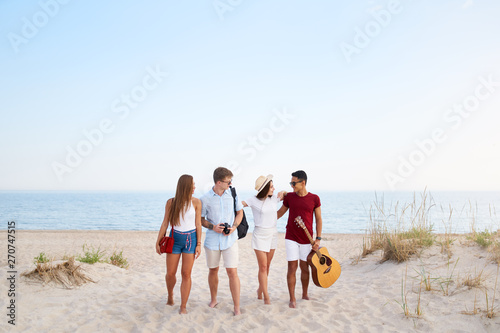 Group of friends going on sandy beach at sunset. Young men and women walking on the shore to relax near sea and play guitar. Teenagers on holidays. Boyfriend and girlfriend embrace. Isolated view.