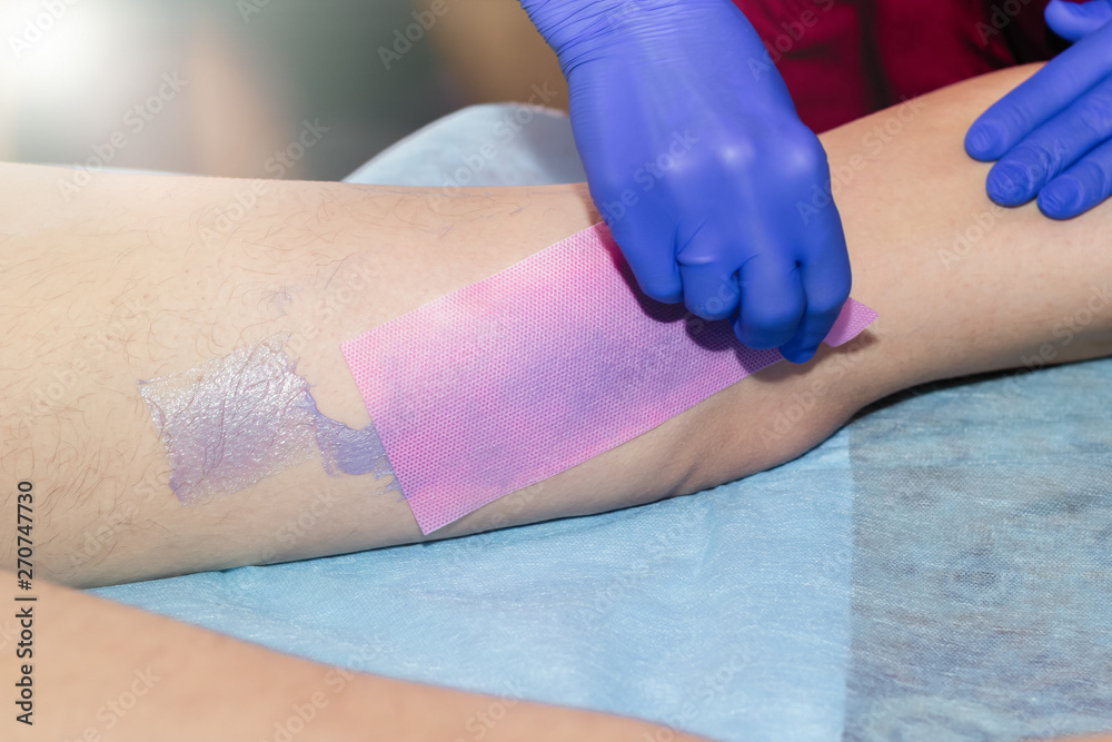Beautician Waxing Leg Of Woman With Wax Strip At Beauty Clinic.