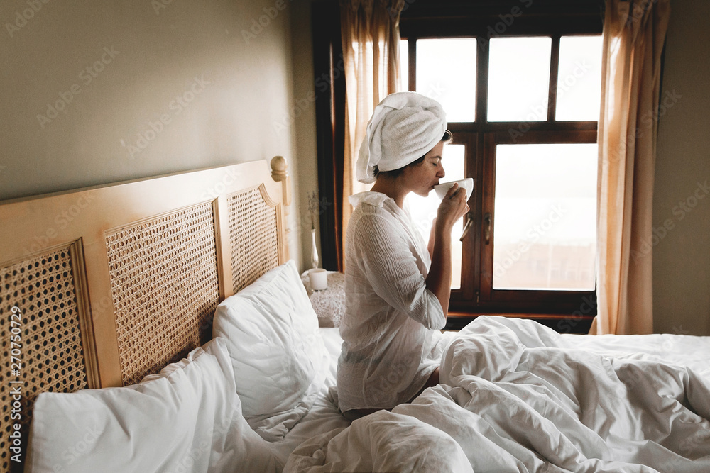 Beautiful happy young woman drinking coffee or in bed in hotel room or home bedroom. Stylish brunette girl with white towel enjoying morning in soft light from window Beautiful happy young woman drinking coffee or in bed in hotel room or home bedroom. Stylish brunette girl with white towel enjoying morning in soft light from window