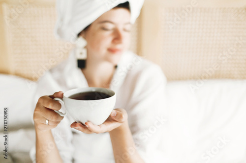 White cup with coffee or tea in girls hands closeup. Beautiful happy young woman drinking tea in bed in hotel room or home bedroom, enjoying morning in soft light. Space for text