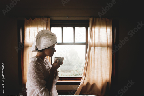 Beautiful happy young woman drinking coffee or tea in bed in hotel room or home bedroom. Stylish brunette girl with white towel enjoying morning in soft light at window. Space for text