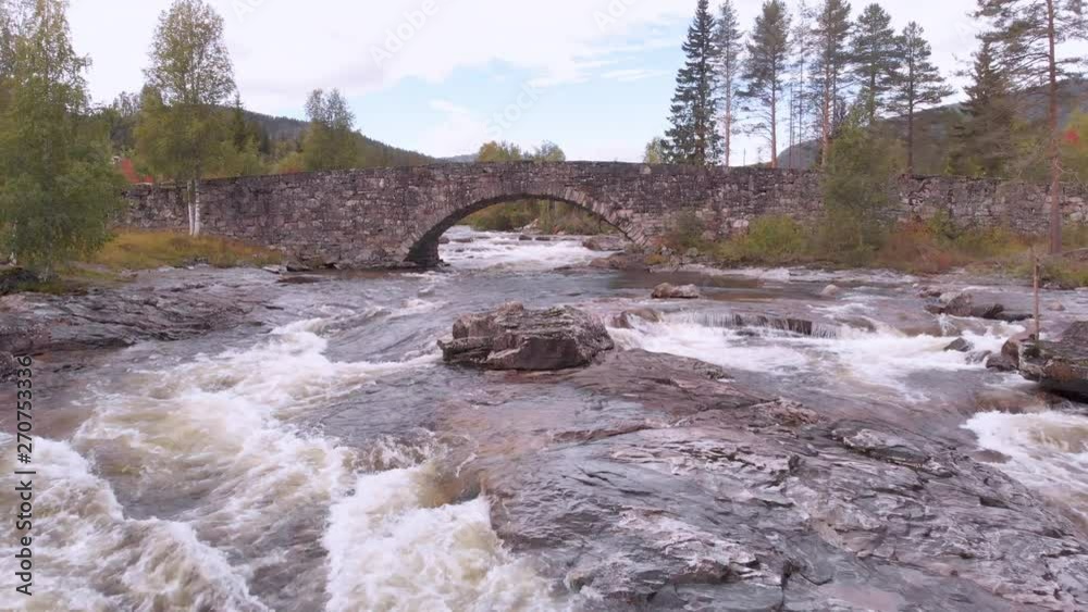 From white water rapids, towards and under a small old stone bridge with an arch. Surrounded by pine and deciduous trees. From white water rapids, towards and under a small old stone bridge with an arch. Surrounded by pine and deciduous trees.