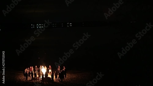 Dark night a few dozen people stand around brightly burning bonfire on the beach photo