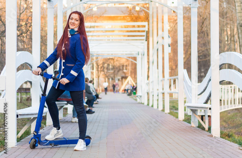 One young caucasian woman with red hair in a blue coat on a blue electric scooter in the park. Eco-friendly transport. Have fun at the weekend.