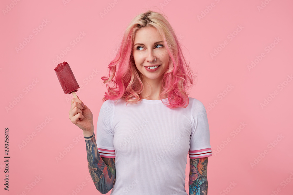Portrait of happy cute girl with pink hair and tattooed hands, looking at ice cream in his left hand and smiling, standing over pink background, wearing a white t-shirt. Portrait of happy cute girl with pink hair and tattooed hands, looking at ice cream in his left hand and smiling, standing over pink background, wearing a white t-shirt.