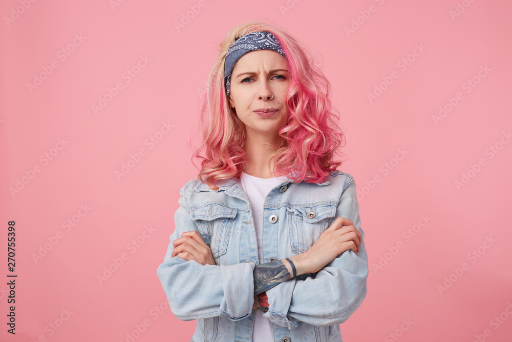 Frowning lady with pink hair and tattooed hand, looking at the camera with disapproval and discontent, standing over pink background with crossed arms, wearing a white t-shirt and denim jacket. Frowning lady with pink hair and tattooed hand, looking at the camera with disapproval and discontent, standing over pink background with crossed arms, wearing a white t-shirt and denim jacket.