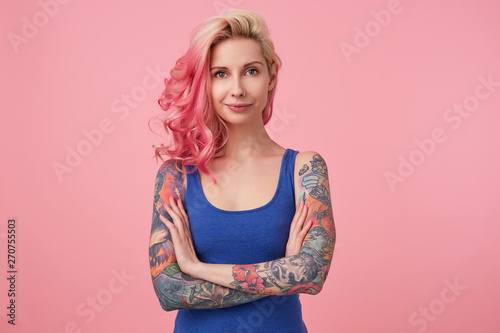 Portrait of positive cute lady with pink hair and tattooed hands, standing over pink background and smiling, wearing a blue shirt. People and emotion concept.
