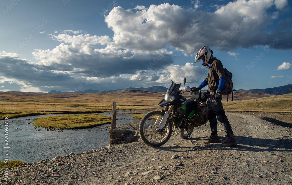 Obraz premium Motorcycle traveler biker man in helmet with suitcases standing on extreme rocky road in a mountain valley on the background of endless steppe