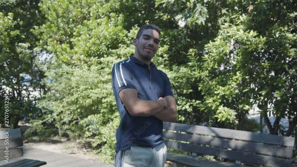 Confident young Caucasian man in dark blue T-shirt standing outside with crossed arms, changing body position, looking at camera, smiling. Lifestyle concept Confident young Caucasian man in dark blue T-shirt standing outside with crossed arms, changing body position, looking at camera, smiling. Lifestyle concept