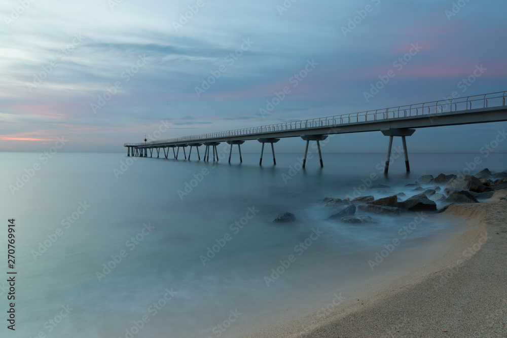 A quiet beach under a cloudy sky at sunrise. Clouds above the pier and at foreground, a long exposure of waves over the rocks and the sand of the beach.