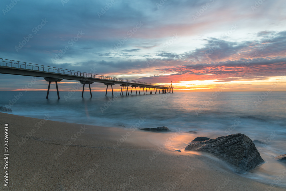 A colored sunrise by the beach. Clouds among the rising sun beyond the dock with silhouettes of pedestrians. At foreground, a long exposure of waves over the rocks of the beach.