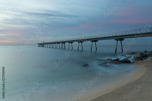 A quiet beach under a cloudy sky at sunrise. Clouds above the pier and at foreground  a long exposure of waves over the rocks and the sand of the beach.