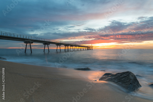 A colored sunrise by the beach. Clouds among the rising sun beyond the dock with silhouettes of pedestrians. At foreground  a long exposure of waves over the rocks of the beach.