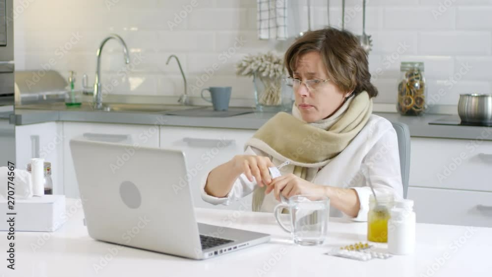 Medium shot of sick woman sitting at kitchen table in front of computer screen, pouring medical powder out of pack into cup of water, mixing it with spoon while talking with her friend by video link