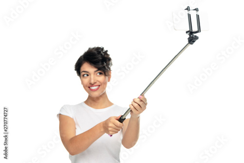 selective focus of happy latin woman holding selfie stick and taking selfie isolated on white