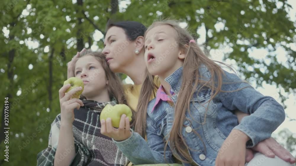 Mature woman sitting under the trees in the park with two cute granddaughters. Girls eating apples looking away. Happy family spending time outdoors. Healthy lifestyle Mature woman sitting under the trees in the park with two cute granddaughters. Girls eating apples looking away. Happy family spending time outdoors. Healthy lifestyle