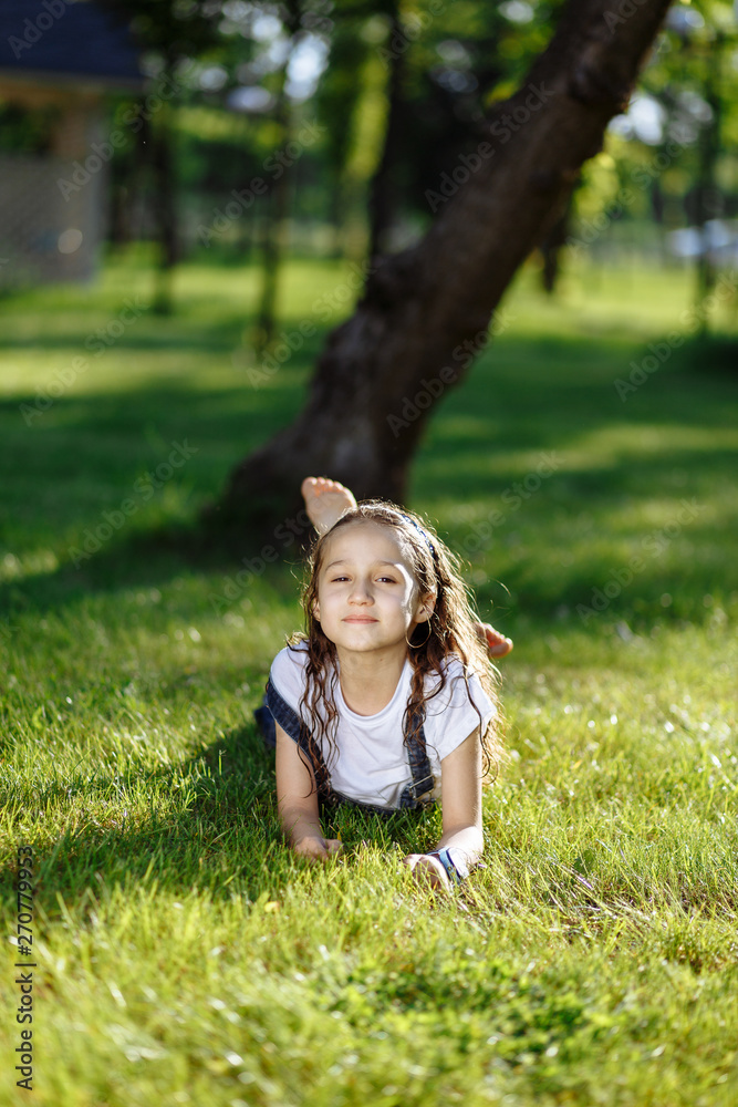 Cheerful little girl laying down on green grass and smiling in summer park. Sunset. Space for text Cheerful little girl laying down on green grass and smiling in summer park. Sunset. Space for text