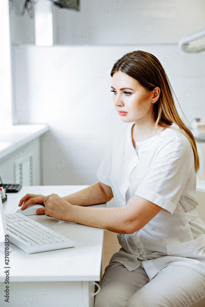 Blonde caucasian woman doctor in white uniform in clinic hospital works on computer and smiles at camera. Blonde caucasian woman doctor in white uniform in clinic hospital works on computer and smiles at camera.