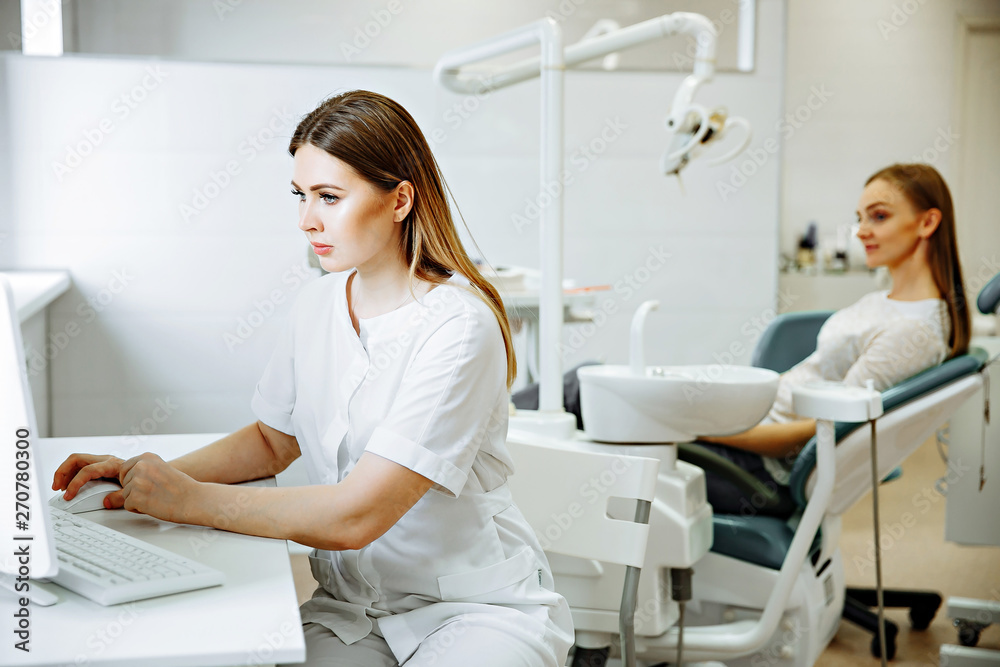 Blonde caucasian woman doctor in white uniform in clinic hospital works on computer and smiles at camera. Blonde caucasian woman doctor in white uniform in clinic hospital works on computer and smiles at camera.
