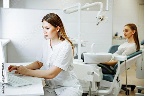 Blonde caucasian woman doctor in white uniform in clinic hospital works on computer and smiles at camera.