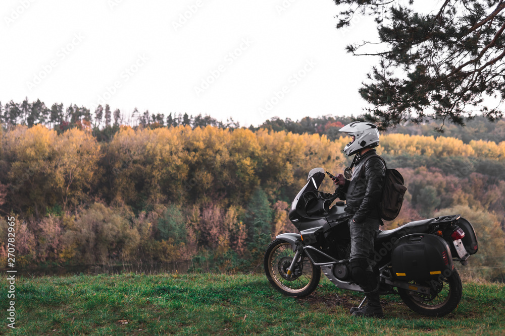 Obraz premium Rider standing with adventure motorcycle, Motorcyclist gear, A motorbike driver looks, concept of active lifestyle, enduro travel road trip. Evening, autumn. Tourist traveler. copy space