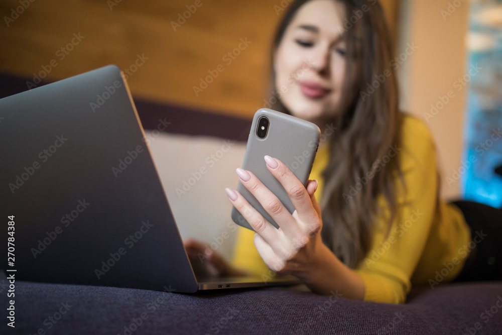 Beautiful young woman in casual clothes is using a smart phone and a laptop, and smiling while lying on couch at home