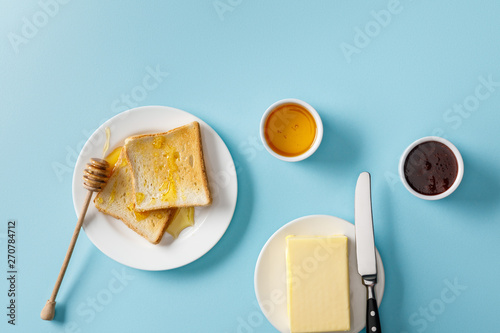 top view of butter, knife on plate, jam, bowls, toasts with honey and wooden dipper on white plates on blue background