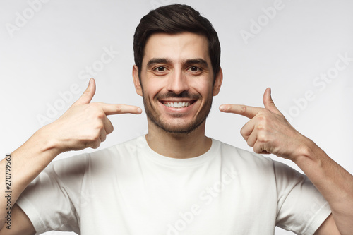 Portrait of smiling young man pointing to his perfect natural white teeth after whitening treatment. Dental clinic ads concept