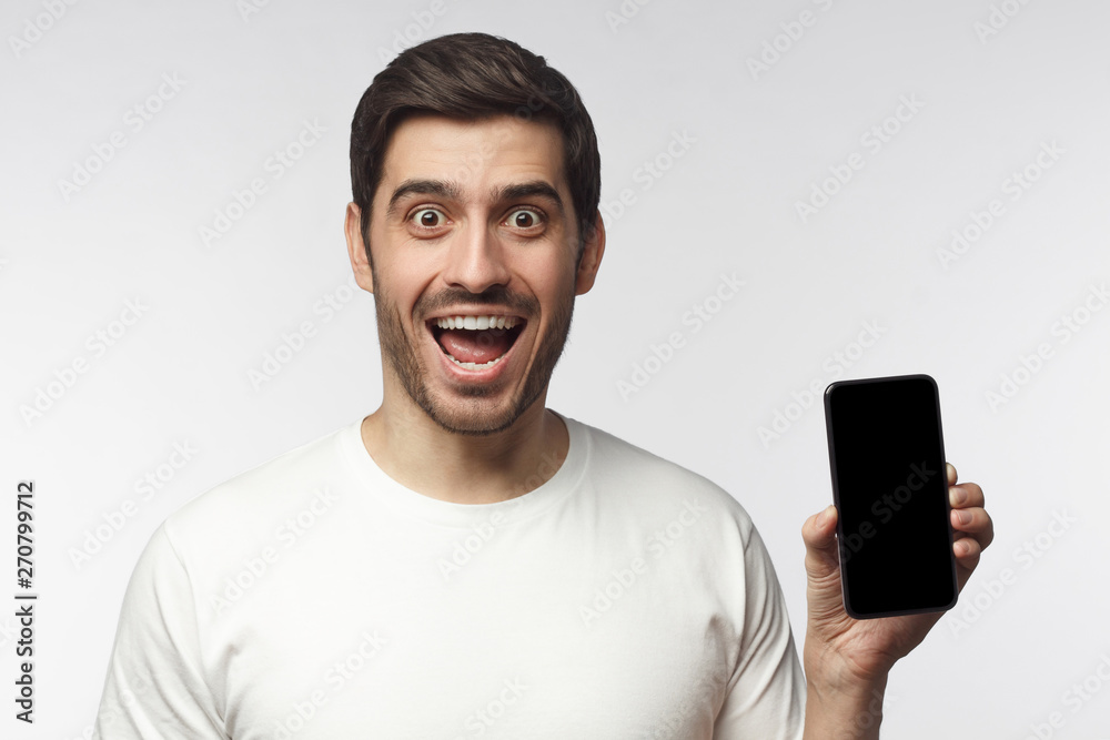 Excited young man holding blank phone with empty black screen, screaming WOW, isolated on gray background Excited young man holding blank phone with empty black screen, screaming WOW, isolated on gray background