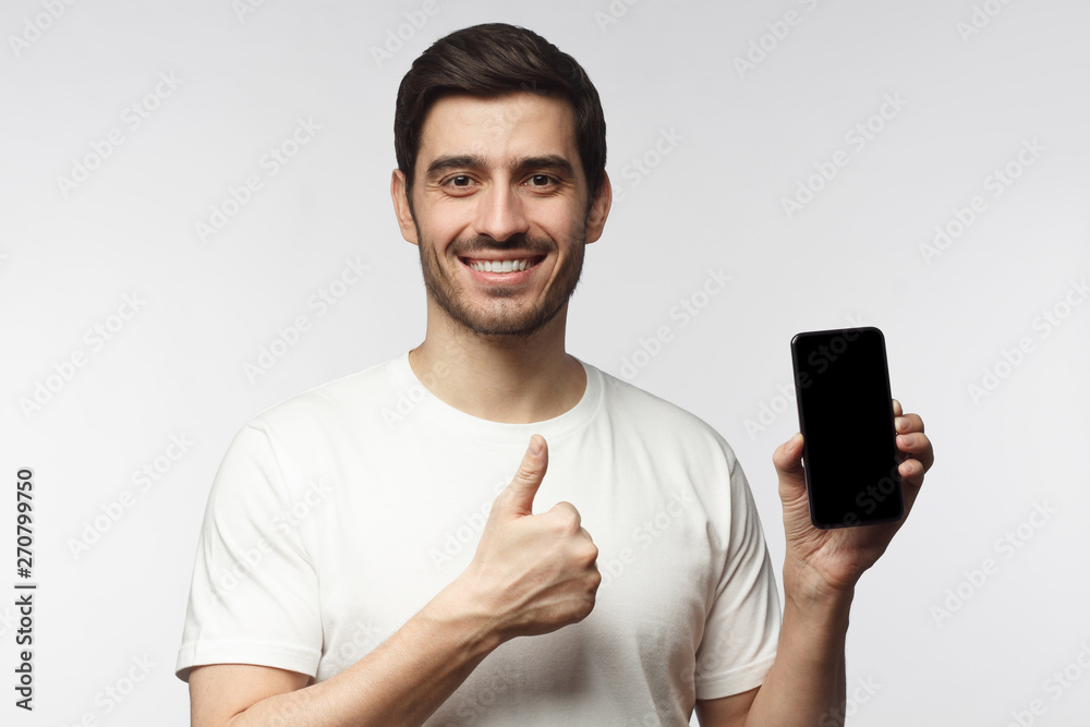 Handsome smiling young man holding blank smartphone, showing thubms up gesture, isolated on gray background Handsome smiling young man holding blank smartphone, showing thubms up gesture, isolated on gray background