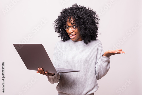 Photo of young african cheerfulwoman standing over grey wall with laptop computer. Looking aside make ask gesture. photo