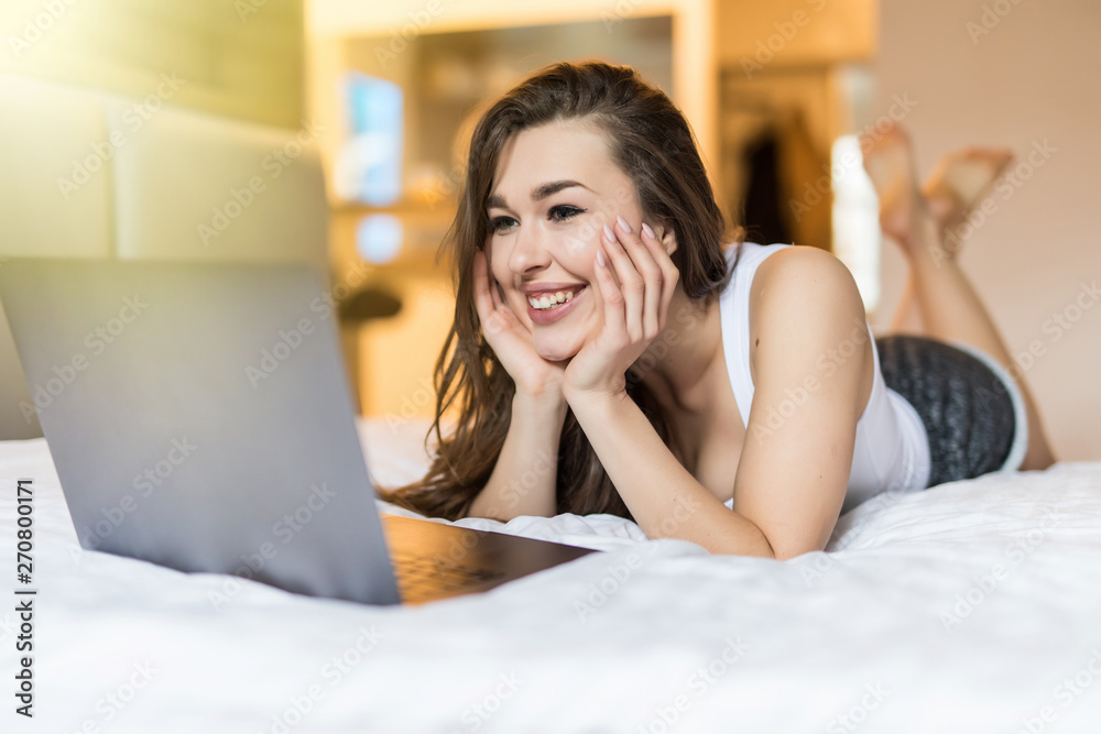 Smiling woman lying down the bed in front of her laptop with her legs raised slightly Smiling woman lying down the bed in front of her laptop with her legs raised slightly
