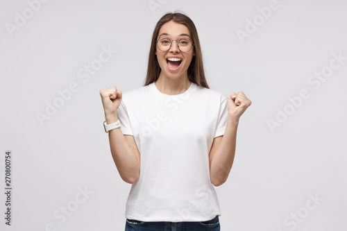 Pretty girl celebrating victory and acting as if she is winner, squeezing fists inl expression of happiness and luck, shouting, isolated on gray background photo