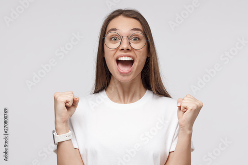 Young attractive woman shouting while her team win, raised both fist in victory gesture, isolated on gray background. Soccer fan concept © Damir Khabirov