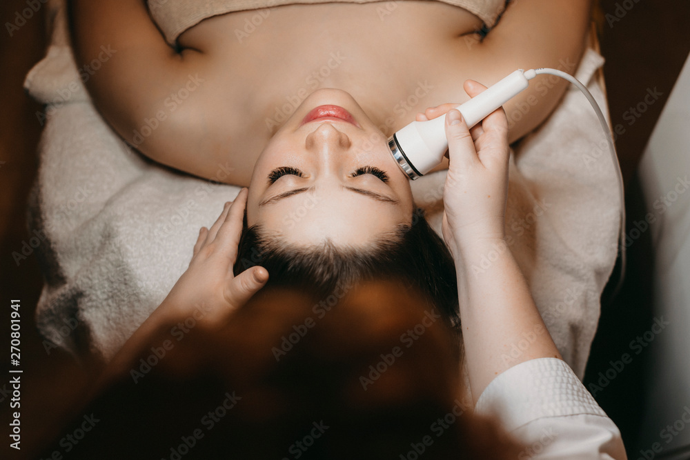 Obraz premium Upper view portrait of a beautiful young female doing a face cleaning with electroporation therapy by a cosmetician.